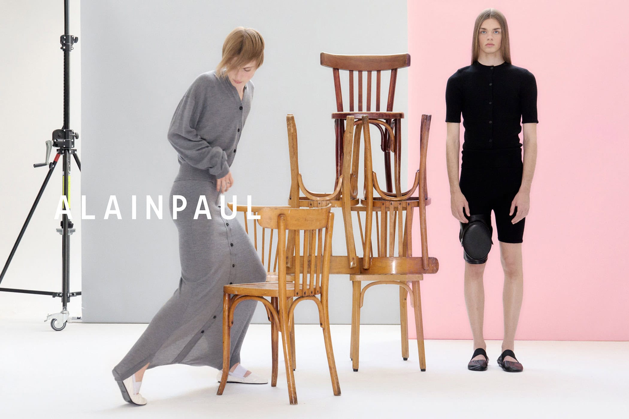 Two models in a studio with stacked wooden chairs; one in a grey knit skirt set and another in a black top and shorts.