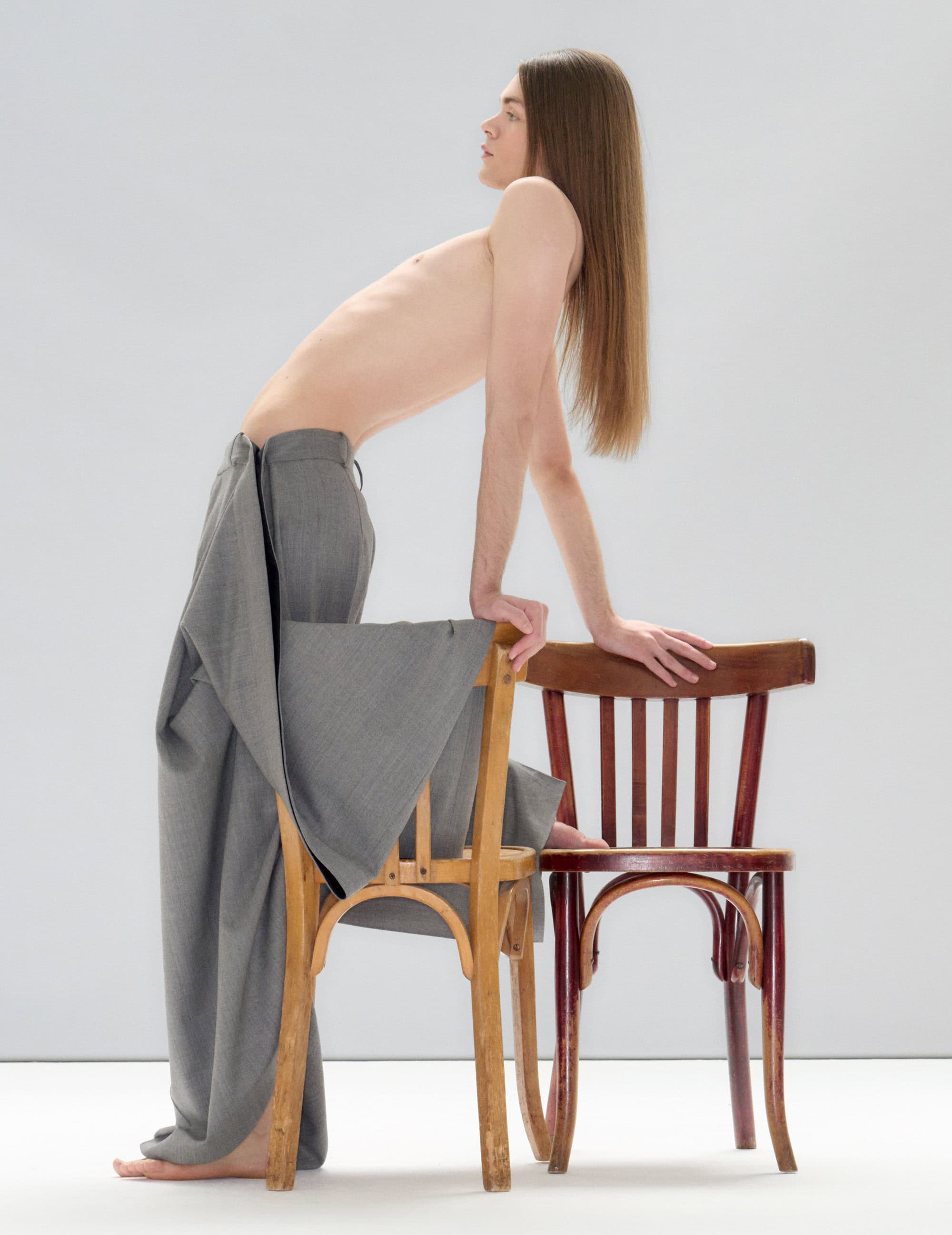 Model with long brown hair wearing grey trousers leaning on two wooden chairs against a plain white studio backdrop.