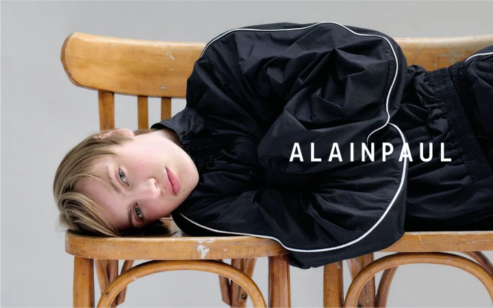 Model lying on a wooden bench wearing a black jacket with white piping against a plain background
