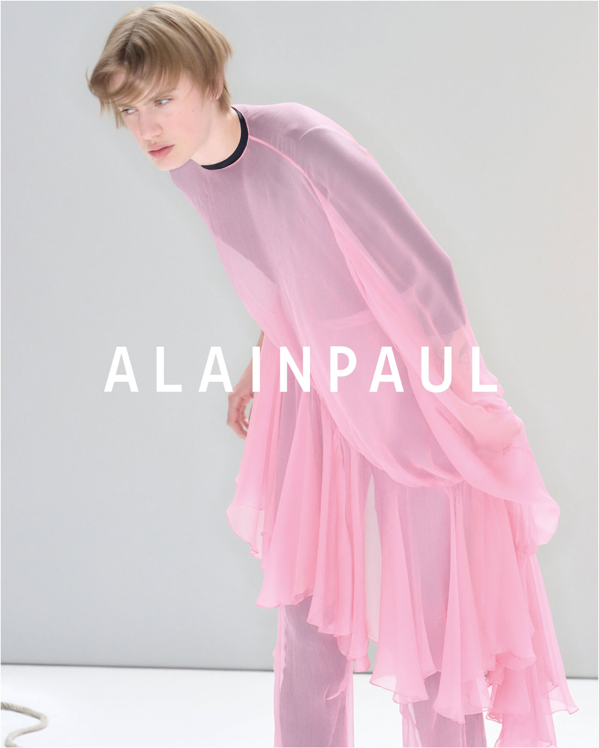 Model wearing a sheer pink tunic leaning forward against a plain light grey backdrop for Alainpaul.