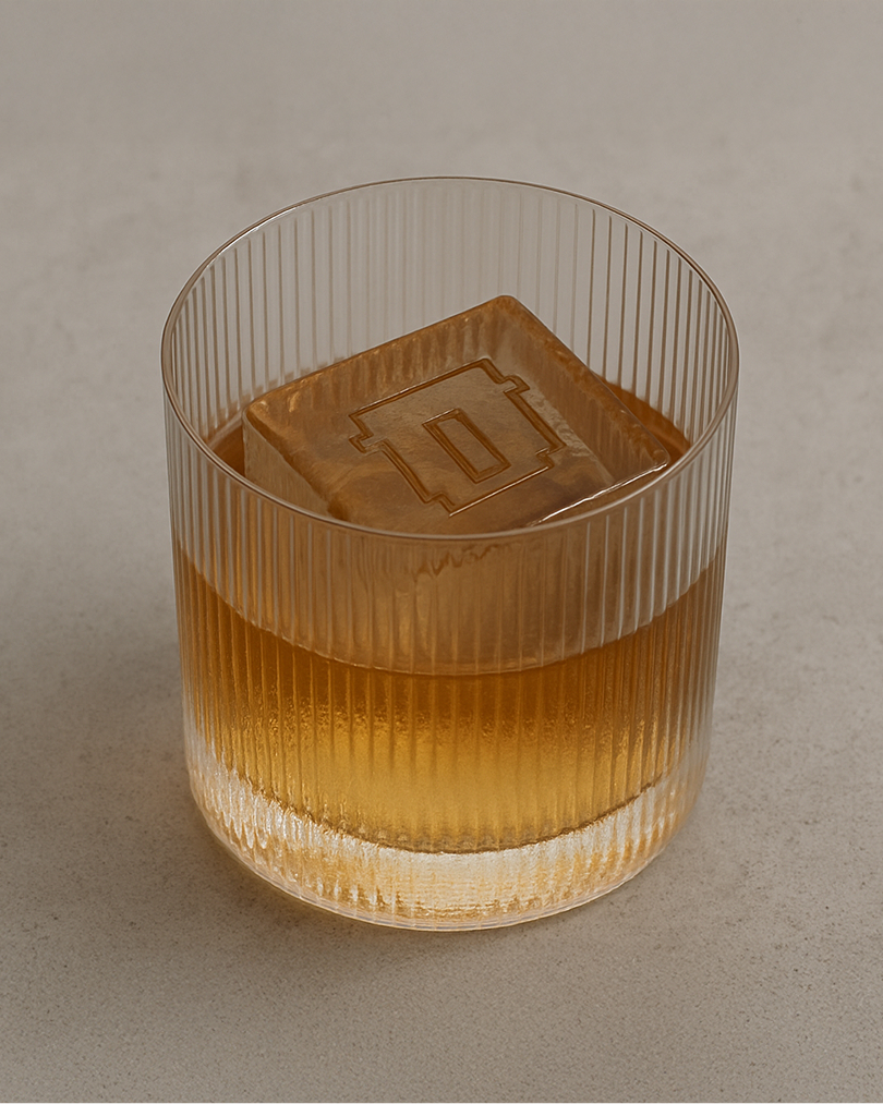 Ribbed glass containing amber spirit with a square ice cube embossed with a logo on a pale grey surface