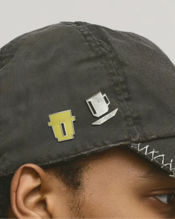 Close-up of a person wearing a dark grey baseball cap pinned with two small coffee cup shaped metal badges by 3TEMP.