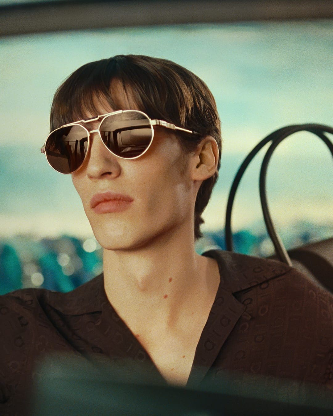 Model wearing gold aviator sunglasses and a dark brown patterned shirt in an outdoor setting for Ferragamo.