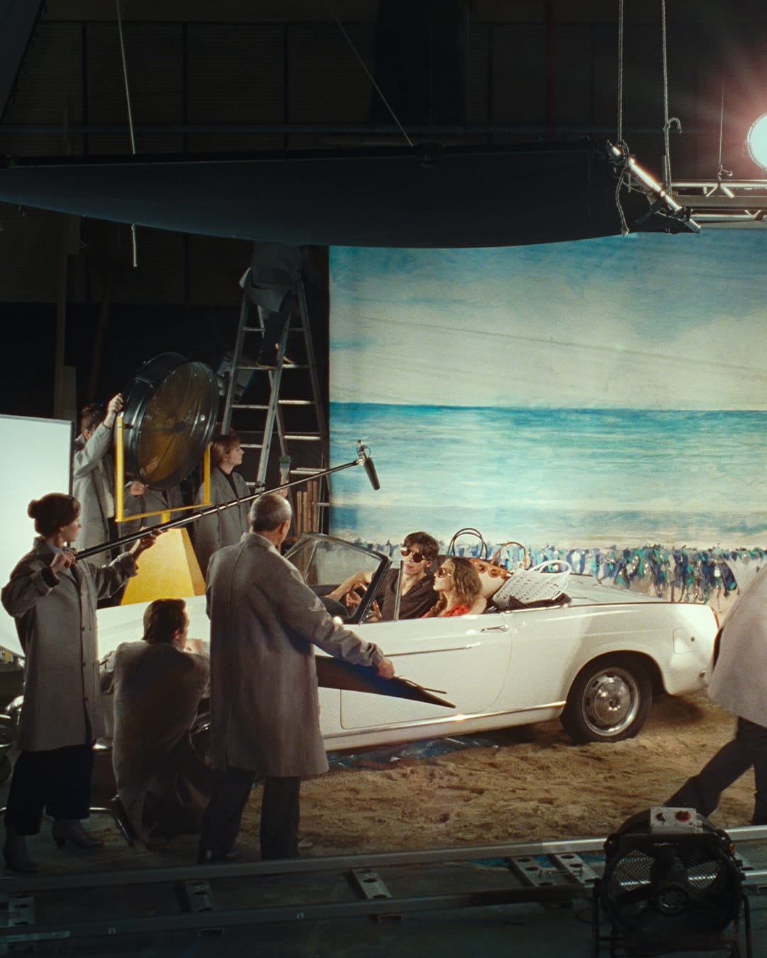 Behind-the-scenes film set with crew members surrounding a white vintage convertible on a soundstage.