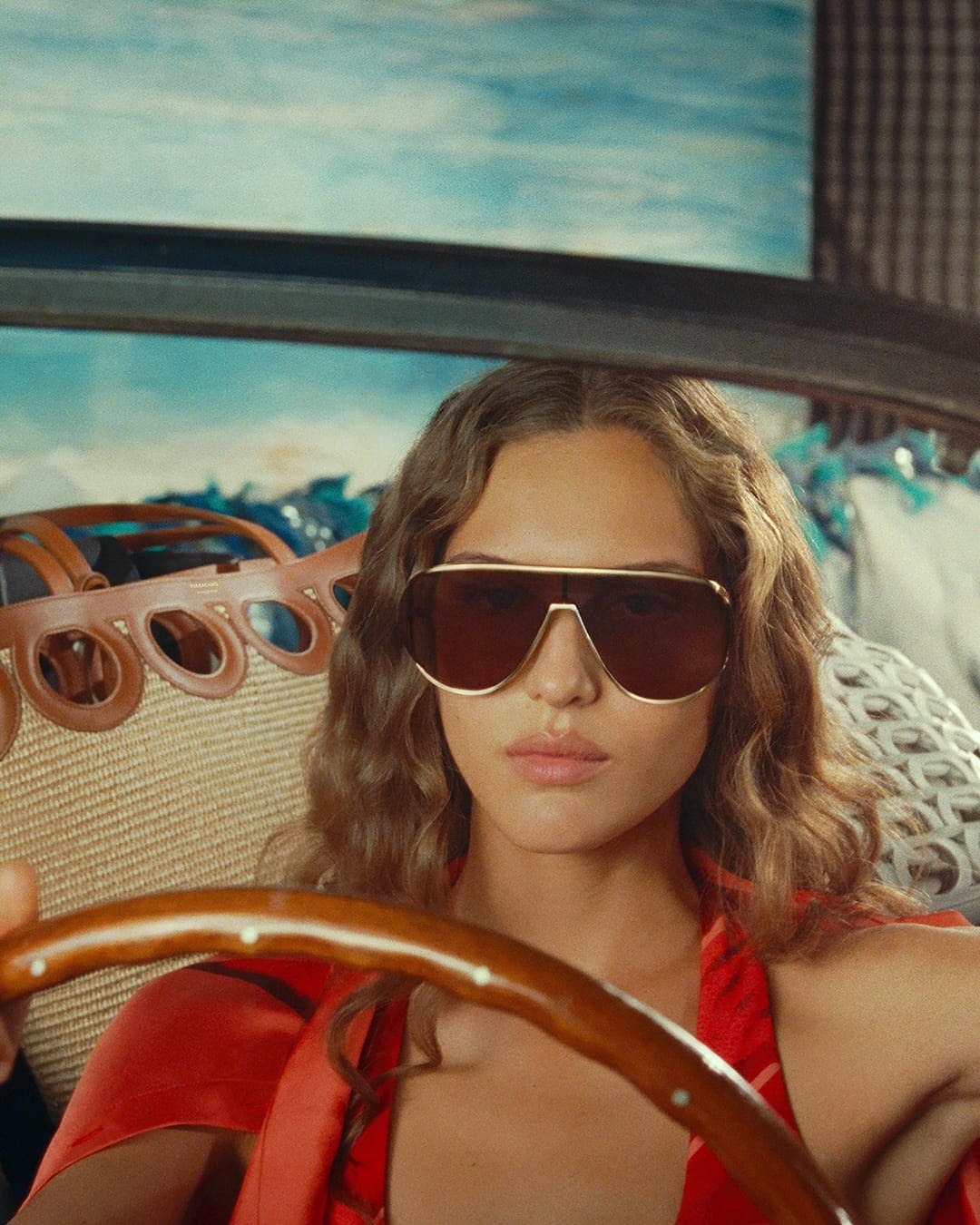 Model Angelina Kendall wearing large sunglasses and a red top in a car for Ferragamo Pre-Fall 2025 campaign