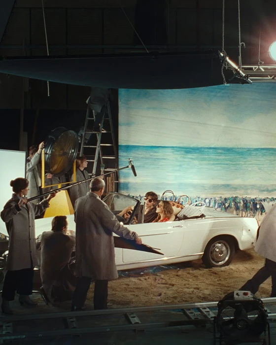 Behind-the-scenes film set with crew members surrounding a white vintage convertible on a soundstage.