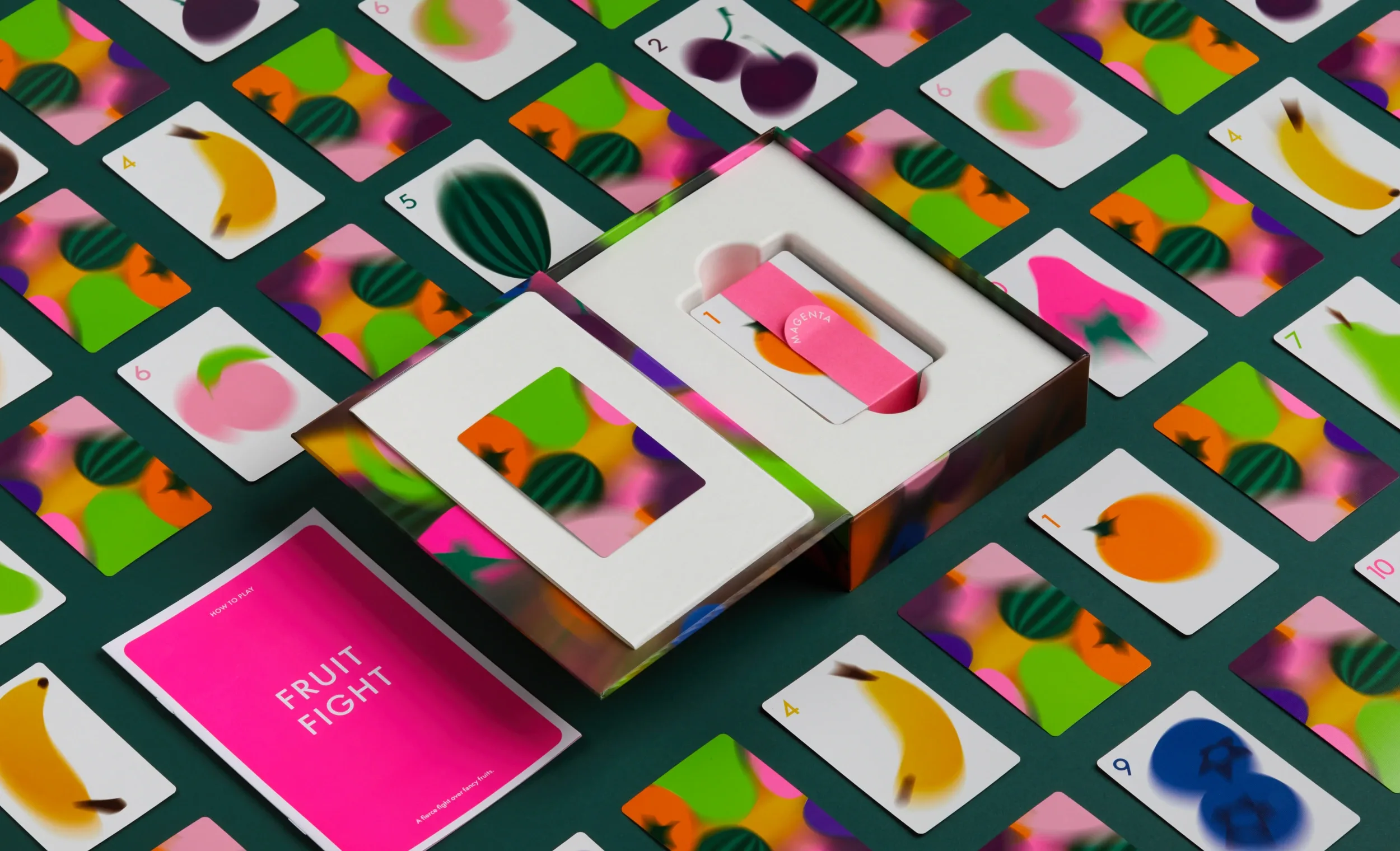 An open board game box surrounded by scattered fruit-themed playing cards on a dark green surface for CMYK brand identity.