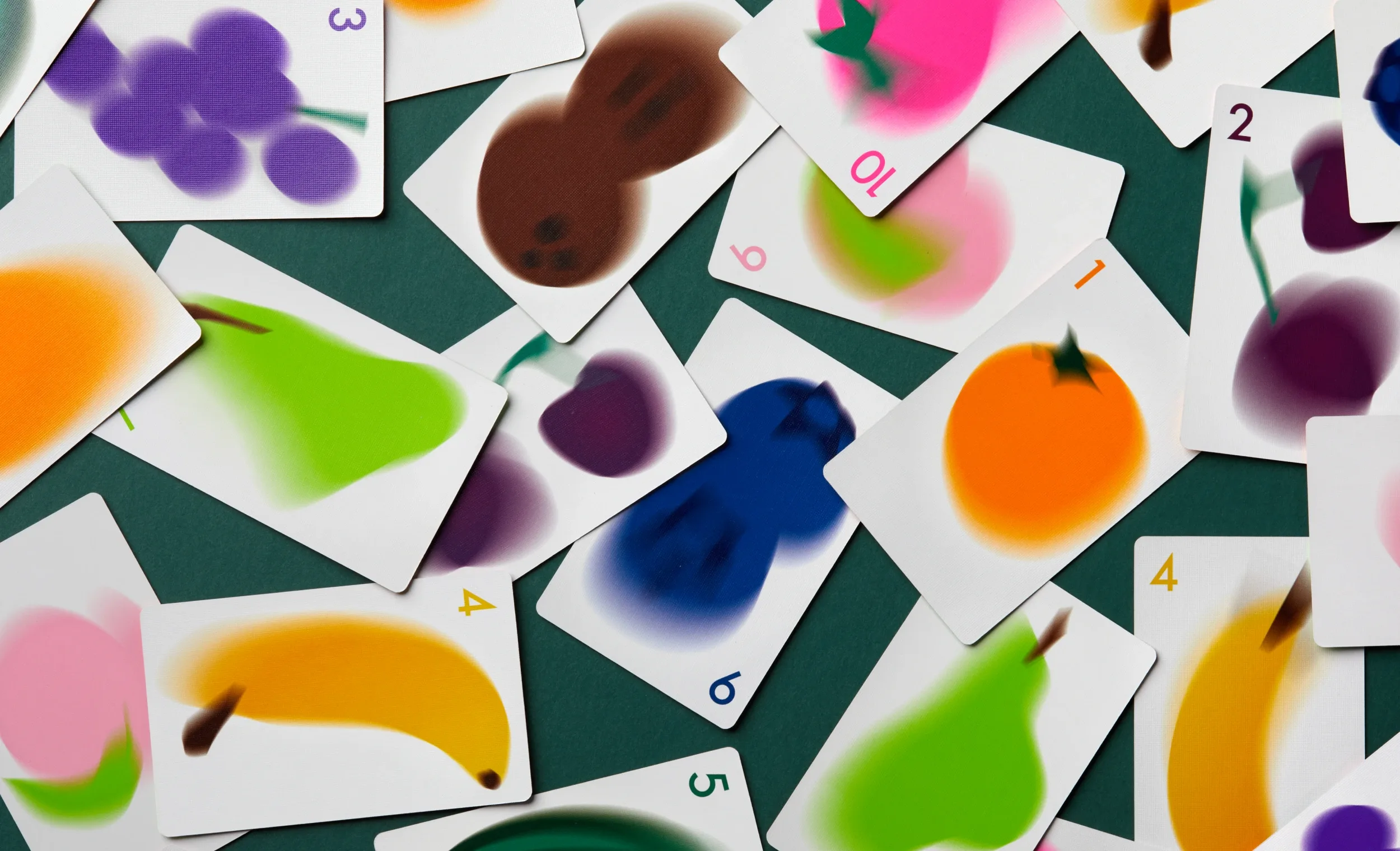 Scattered white playing cards featuring airbrushed fruit illustrations and numbers spread across a dark green surface