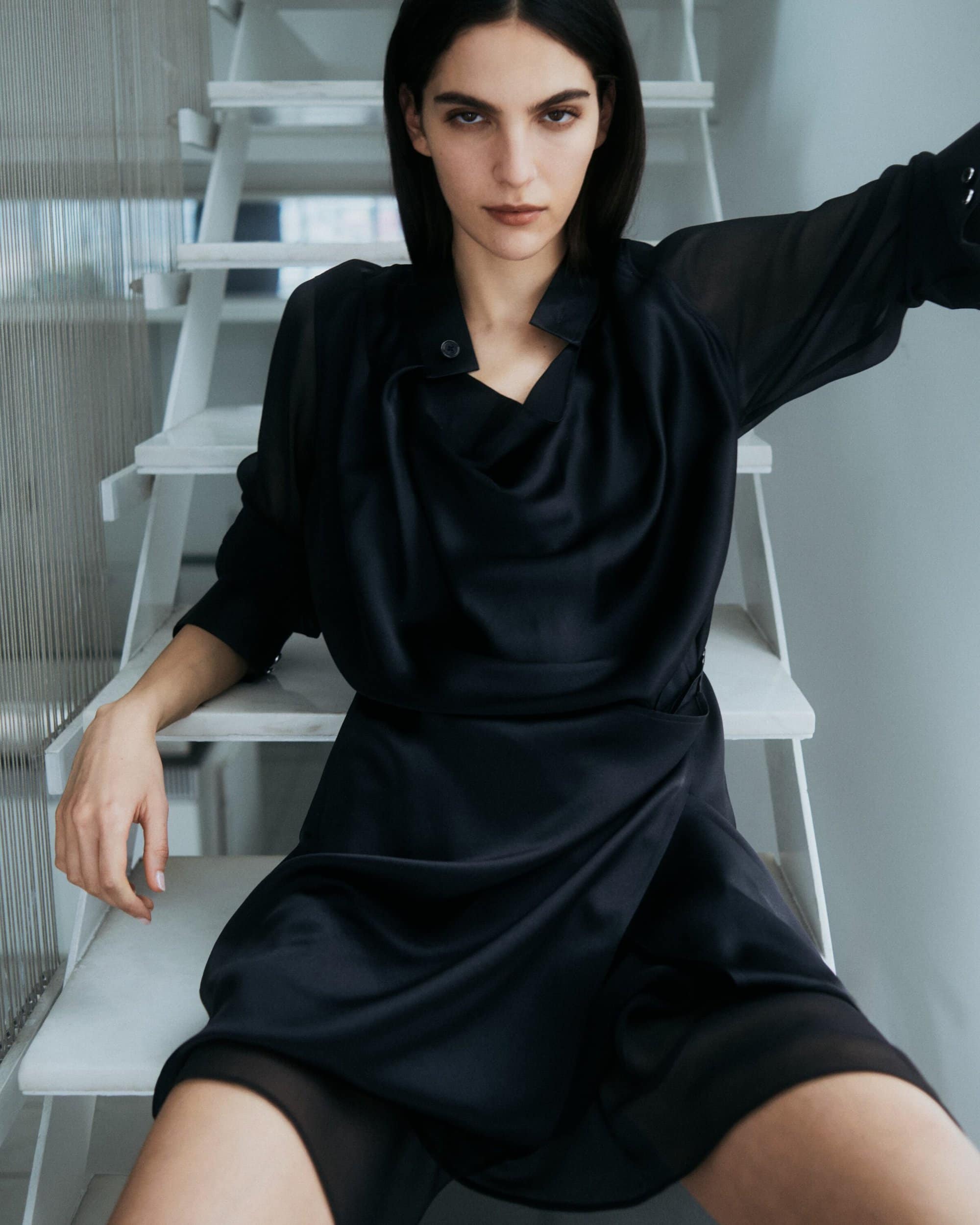 Model wearing a black silk dress seated on white stairs for Helmut Lang pre-fall campaign