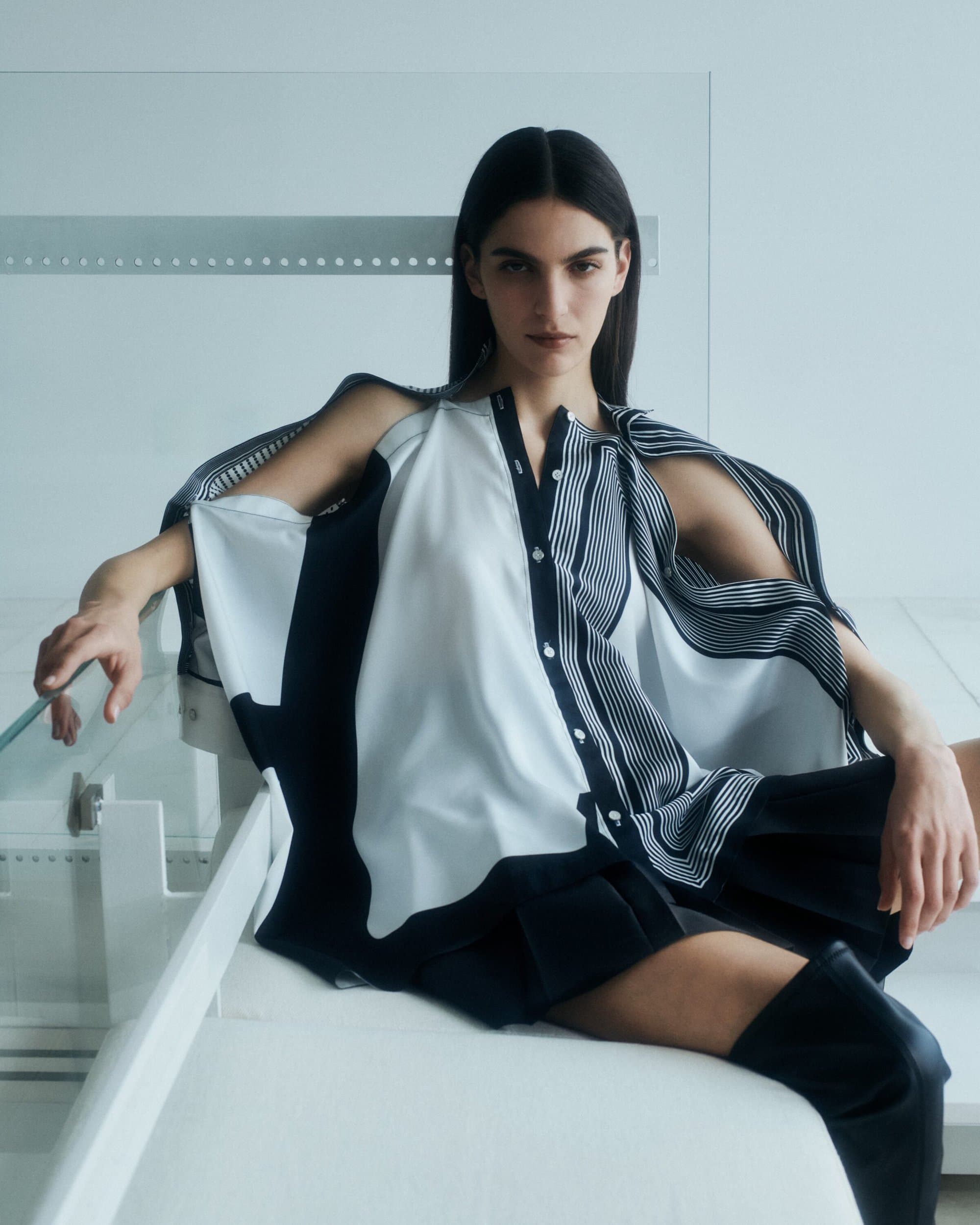 Model seated wearing a black and white patterned shirt and dark boots against a modern white background