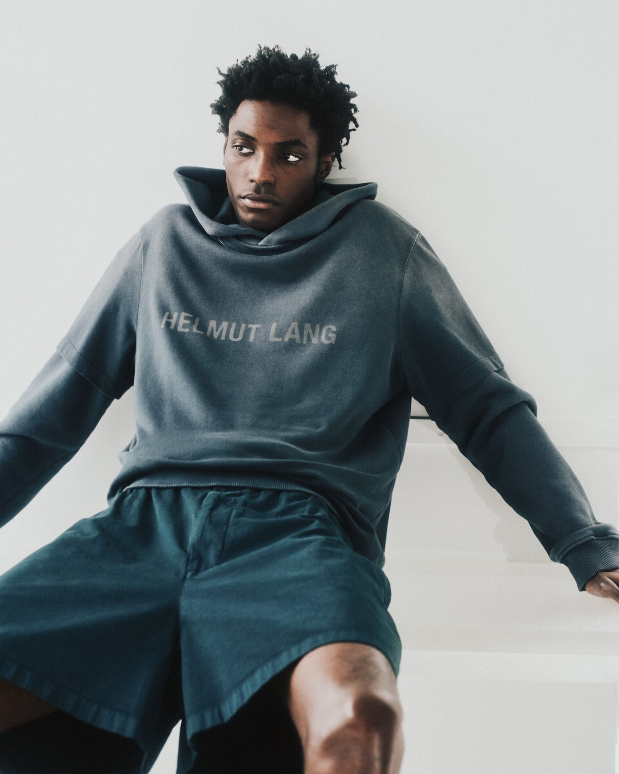 Black man wearing a dark hooded sweatshirt with Helmut Lang branding and matching shorts against a plain white background