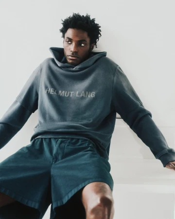 Black man wearing a dark hooded sweatshirt with Helmut Lang branding and matching shorts against a plain white background