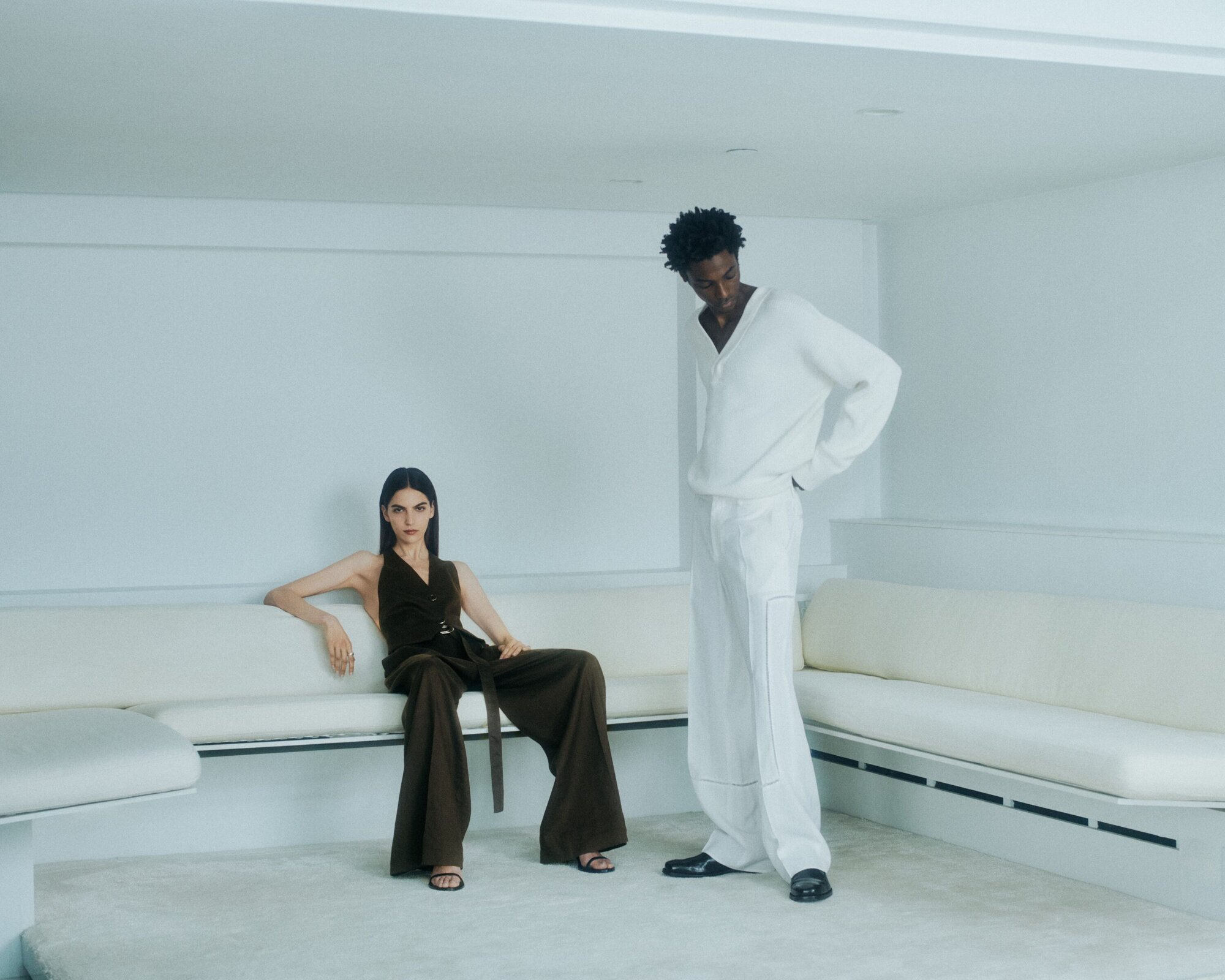 Woman in dark brown trousers seated on a white sofa alongside a man standing in white attire for Helmut Lang.
