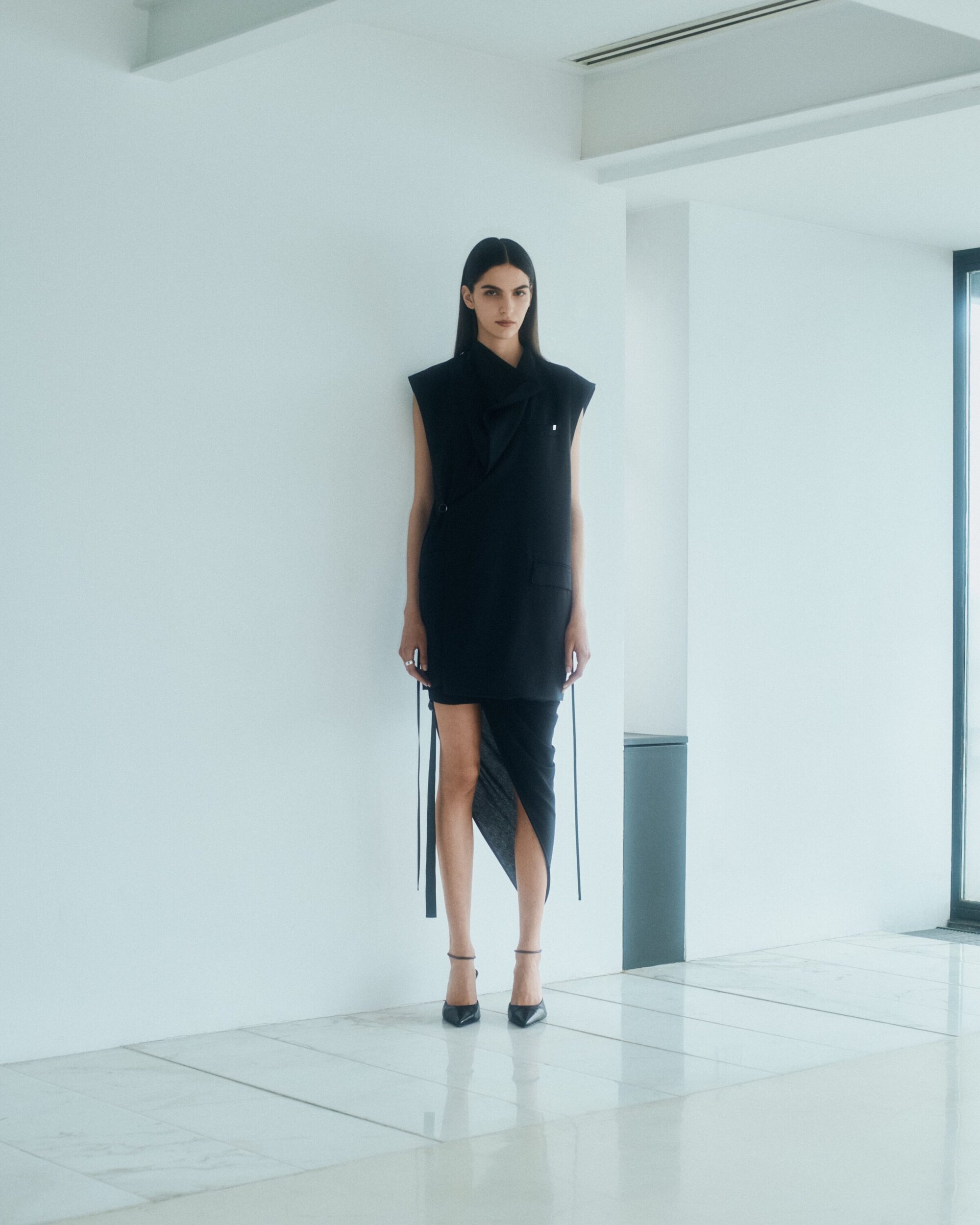 Model standing against a plain white interior wall wearing a black sleeveless dress and dark heeled shoes.