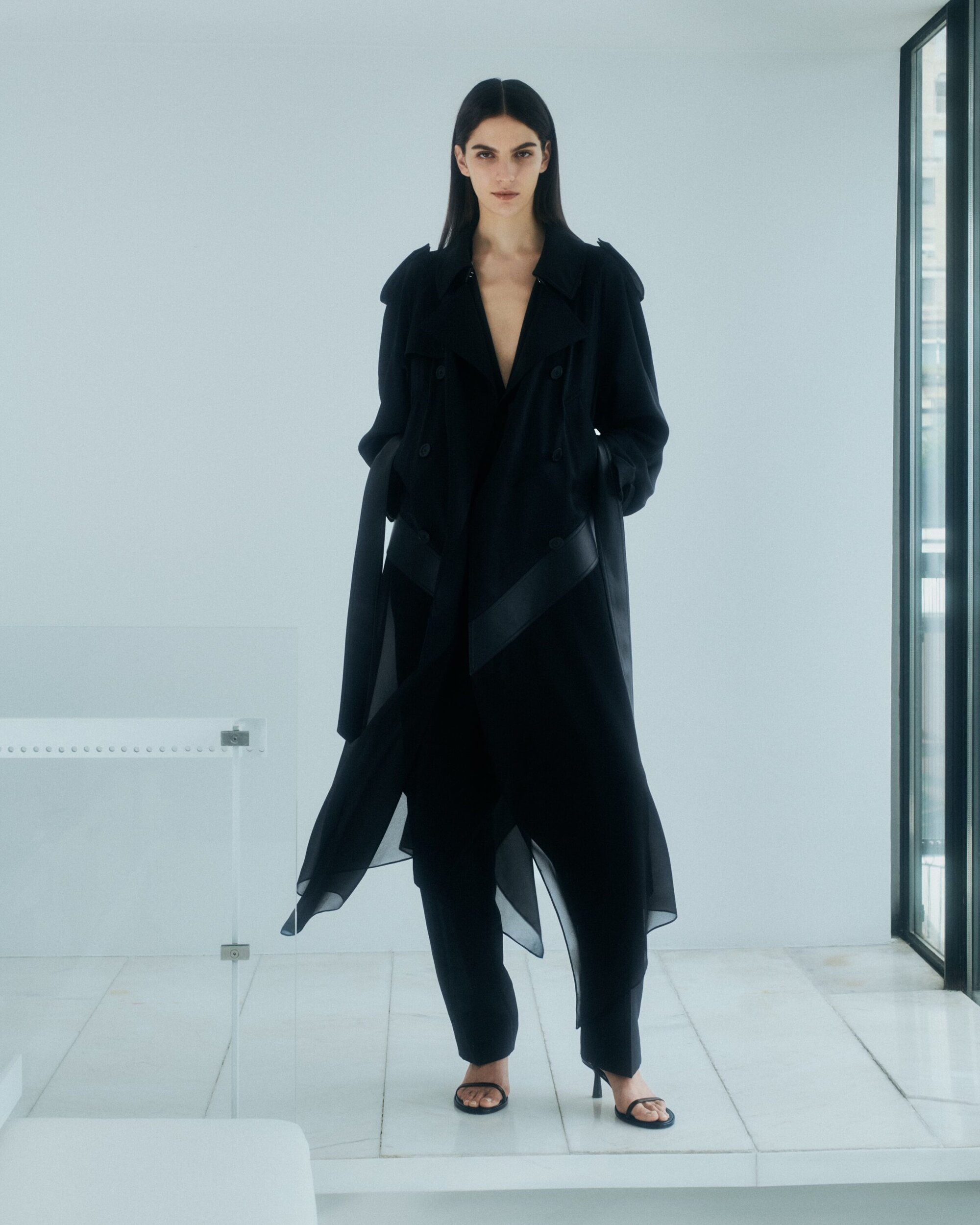 Model in a black coat and trousers standing against a minimal white wall for Helmut Lang pre-Fall 2024 campaign.