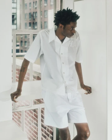 A Black man wearing a white button-down shirt and shorts leaning against a white ledge in an indoor setting.