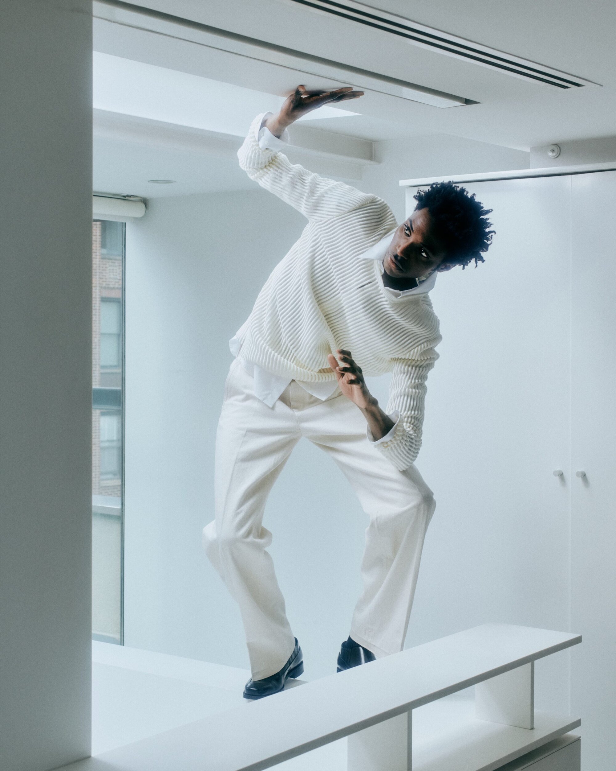 Model in white knit sweater and trousers pressing hands against ceiling in a bright white interior