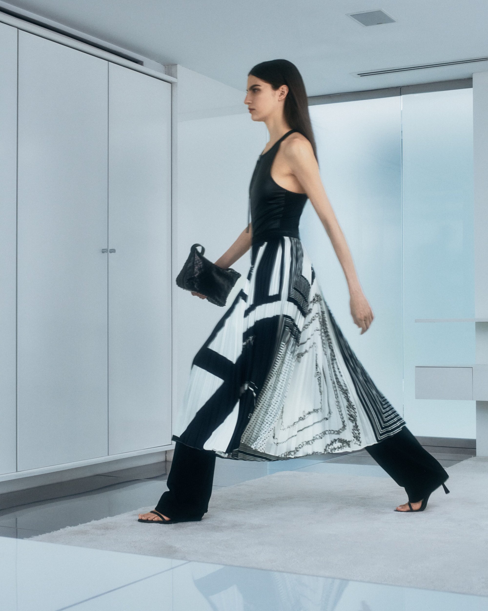 Model walking in a black top and geometric patterned black and white skirt holding a clutch bag