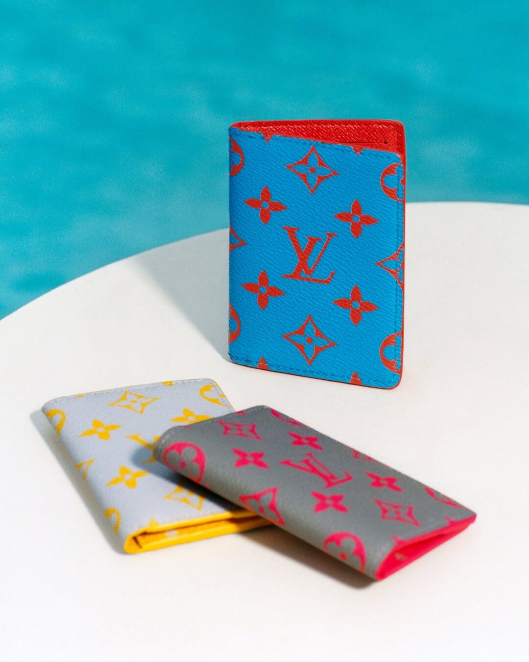 Three colorful designer monogram leather card holders displayed on a white surface with blue water behind.
