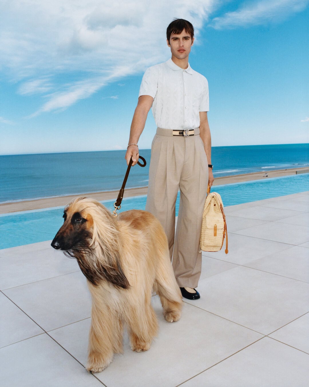 Man in white polo shirt and beige trousers holding an Afghan Hound dog by a leash near a pool and ocean.