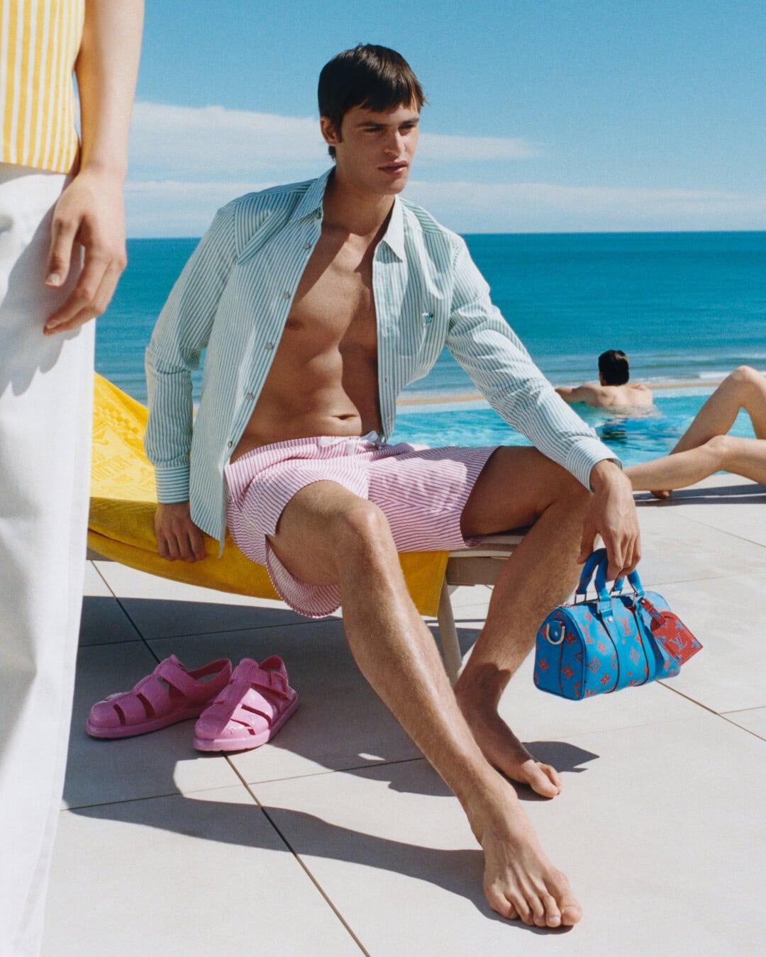 Man seated by a pool wearing striped shirt and pink swim shorts holding a blue handbag, photographed by Rosie Marks for Louis Vuitton.