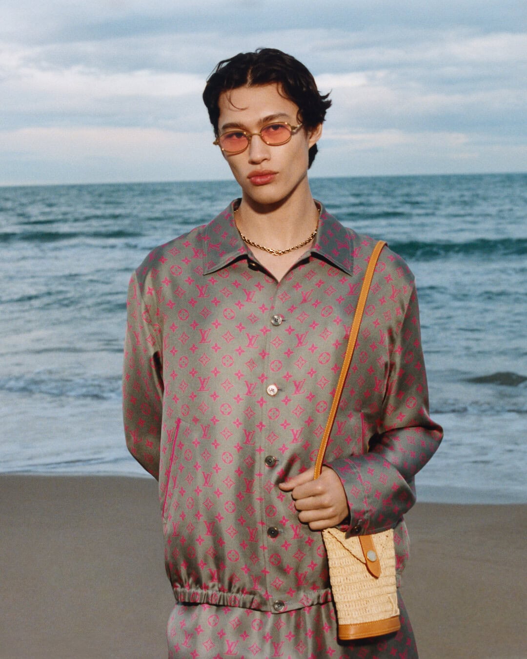Man wearing pink-tinted sunglasses and a patterned button-down shirt standing on a sandy beach by the ocean.