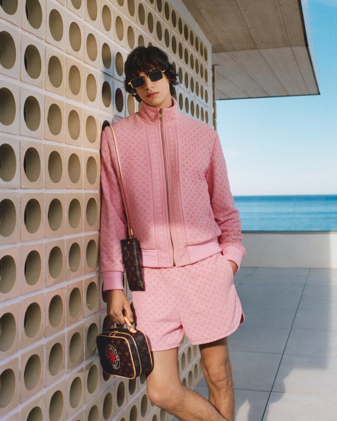 Male model in pink monogram jacket and shorts leaning against an outdoor patterned wall with ocean in background