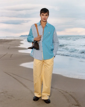 Man wearing a striped shirt and yellow trousers standing on a sandy beach carrying a brown leather bag