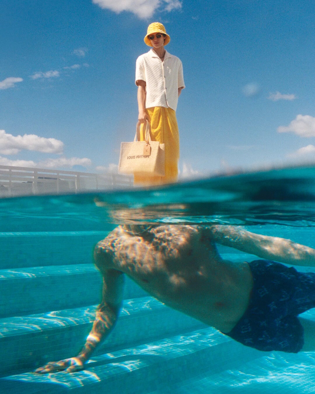 Split-level view of a model standing poolside in yellow shorts and a white shirt holding a canvas tote, with another person swimming underwater.