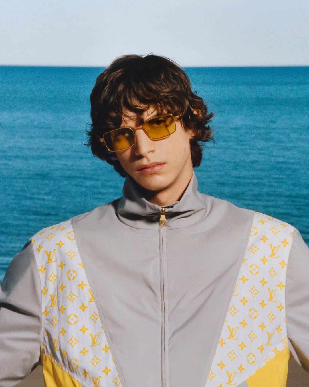 Close-up of a model wearing yellow-tinted sunglasses and a grey zip-up jacket against a blue ocean background.