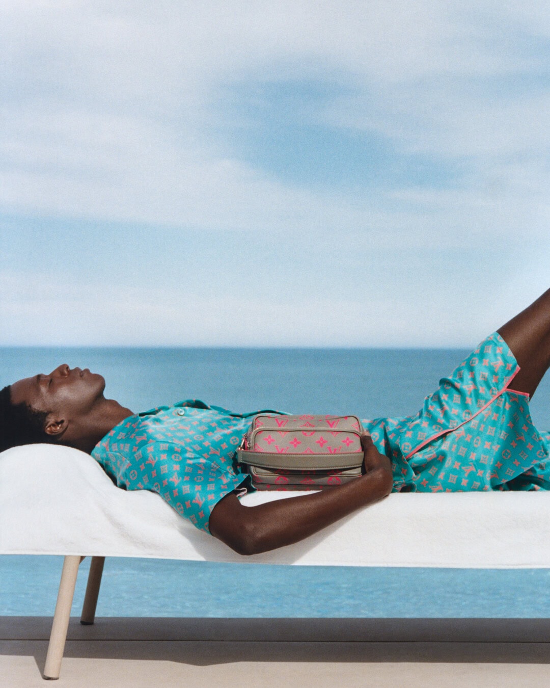 A Black man relaxing on a white sun lounger holding a monogram handbag against an ocean and sky background.
