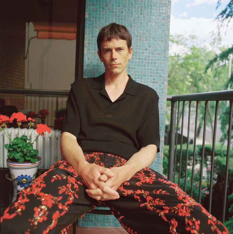 Adult male sits with hands clasped in floral pants on outdoor patio with potted plants and a tree-lined background under a blue sky.
