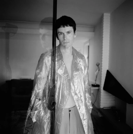 Portrait of a male model in a translucent overcoat, standing next to a black metal post inside a minimally decorated room, black and white.