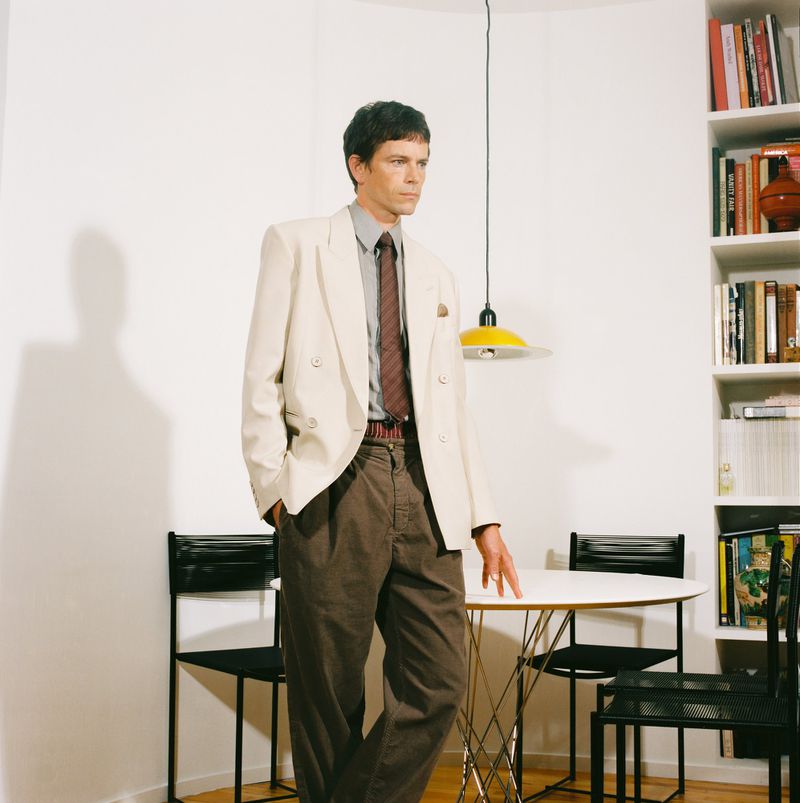 Stylish man in a tan suit poses indoors near a table and chairs with a bookshelf visible. The light is bright and the setting is clean.