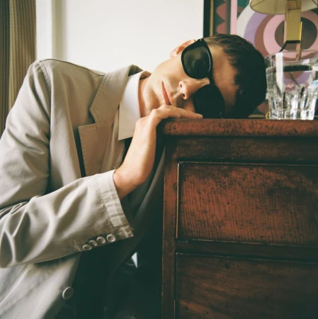 An adult male rests his head on a wooden dresser while wearing a light colored suit and dark sunglasses in an indoor setting.