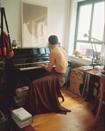 Back view of a person playing a black Yamaha piano in a room with natural light, books, and artwork for a creative, musical scene.