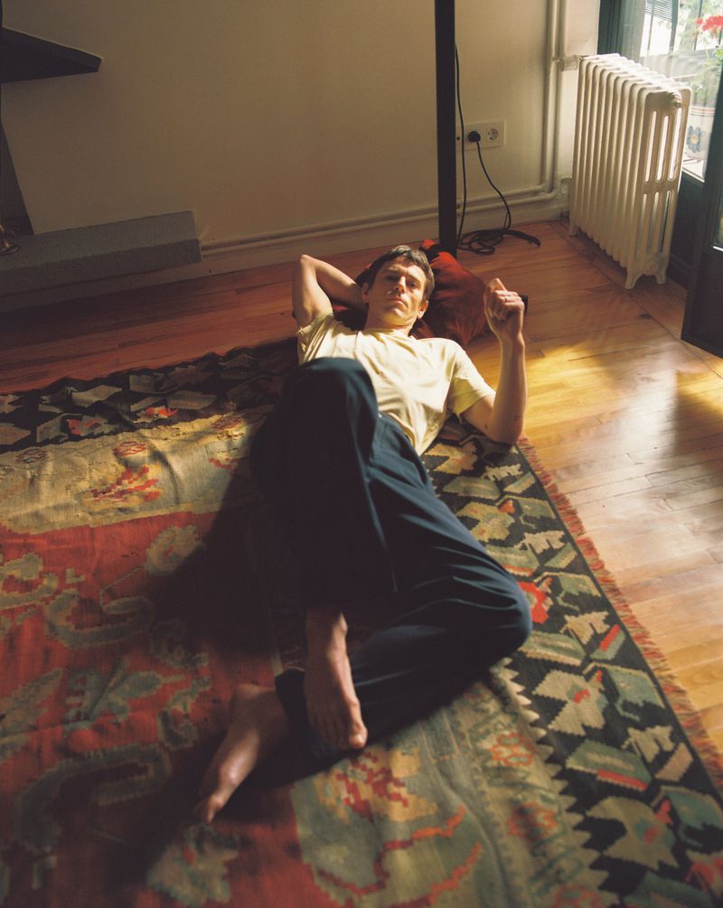 Adult male casually lying on a patterned rug in a domestic setting with wooden floors, natural light, and a radiator in view.