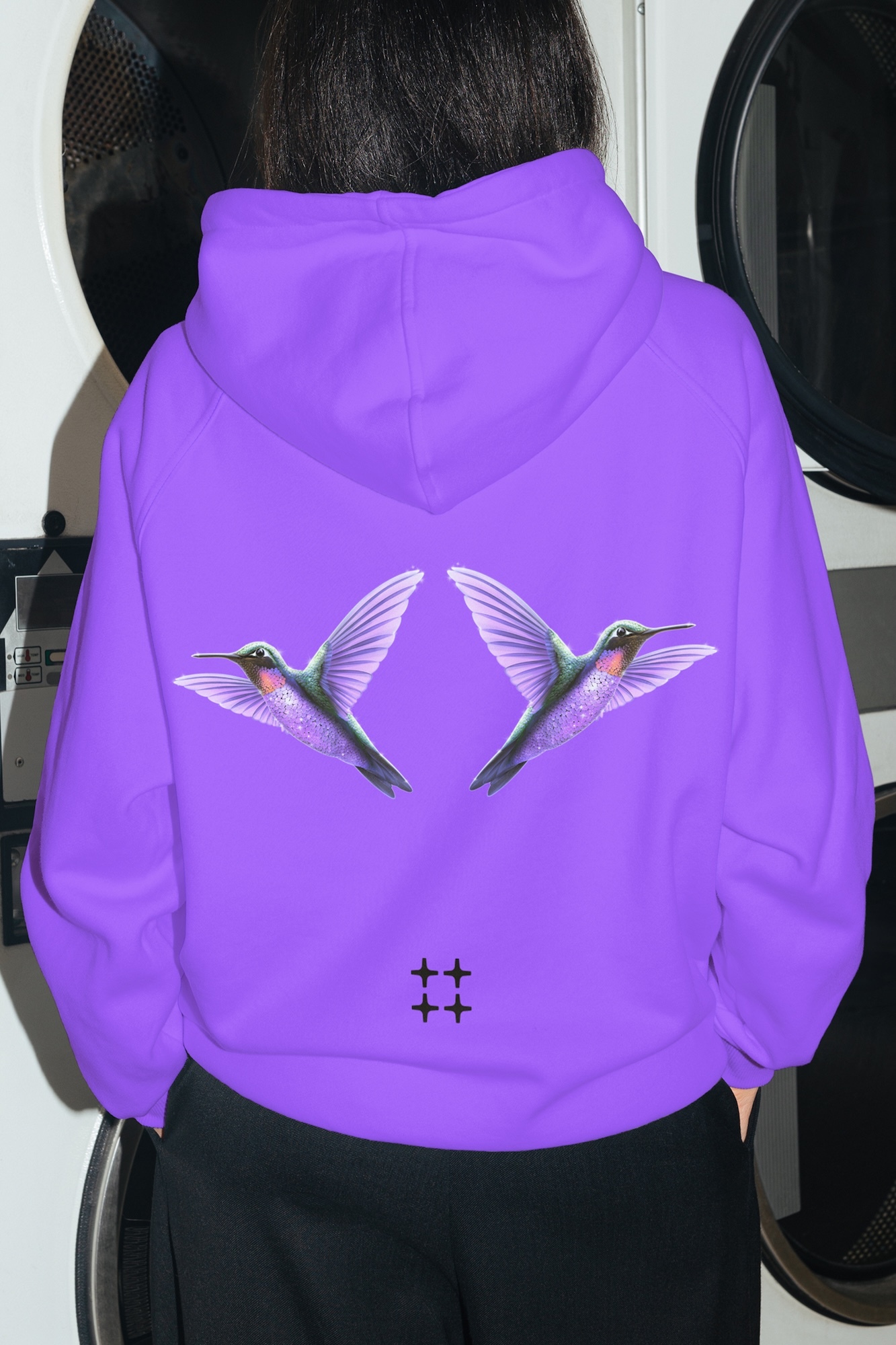 Person wearing a bright purple hoodie with a hummingbird graphic on the back standing in a laundromat
