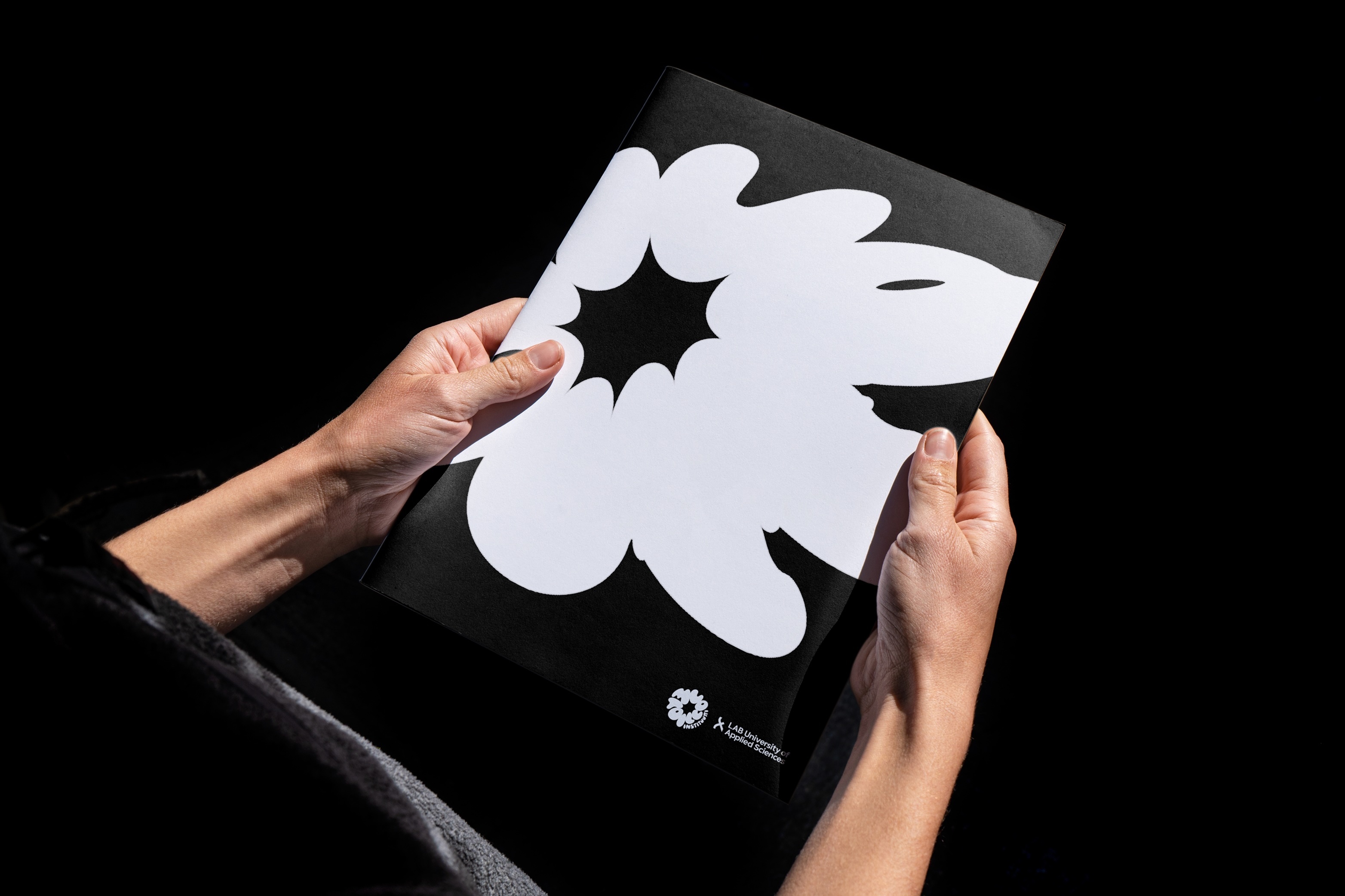 Person holding a black booklet featuring a bold white organic graphic shape against a dark background.