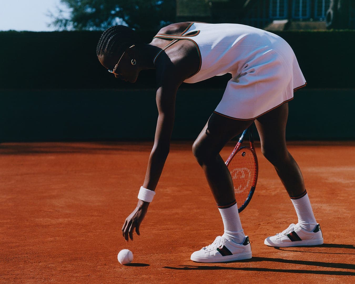 Black female tennis player wearing white sportswear reaching down to pick up a tennis ball on a red clay court