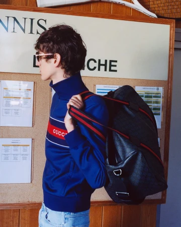 Young man in profile wearing a blue tracksuit jacket and sunglasses carrying a dark duffel bag in front of a bulletin board