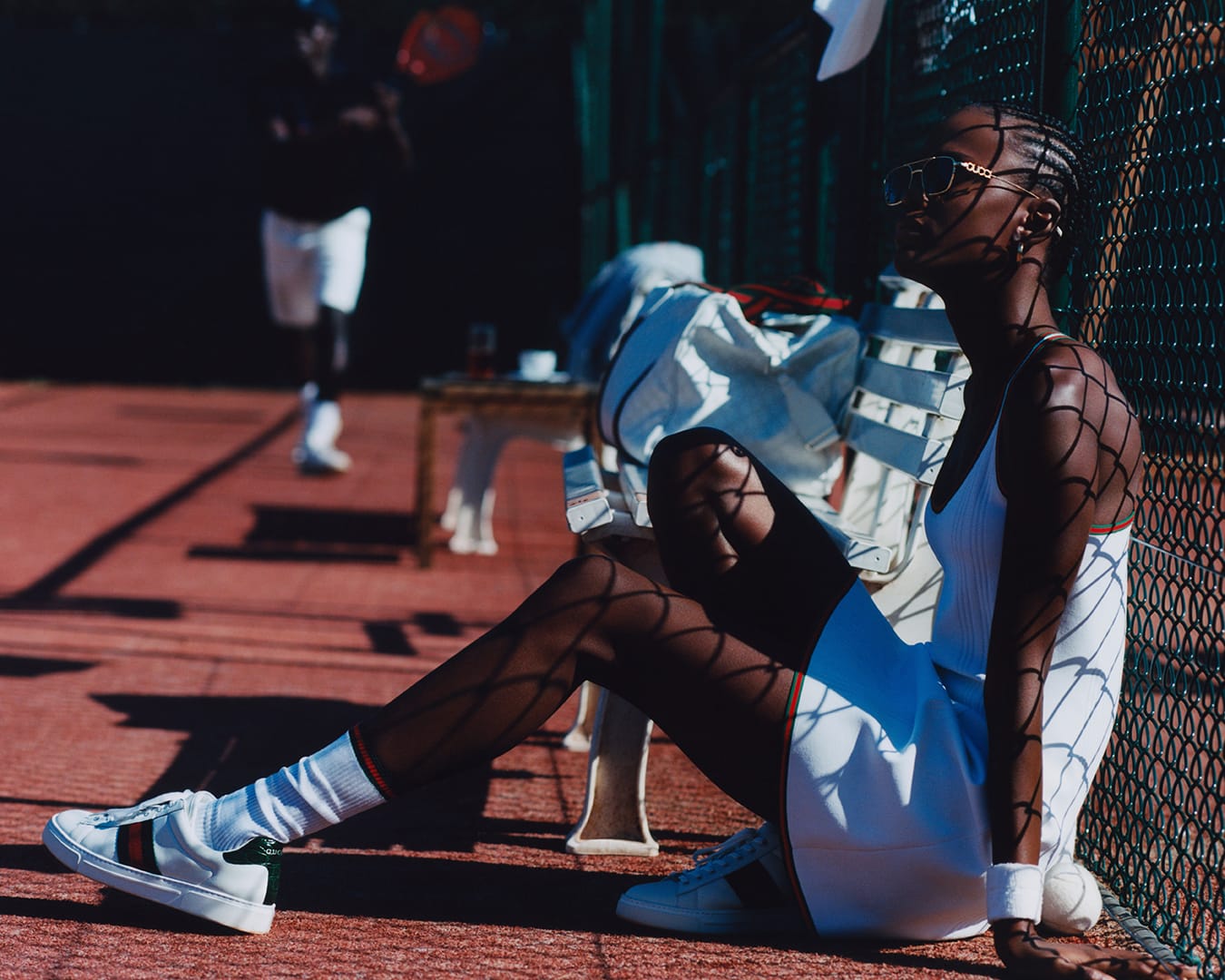 Black woman wearing a white tennis dress and sunglasses seated on a court bench for Gucci Tennis 2025 campaign.