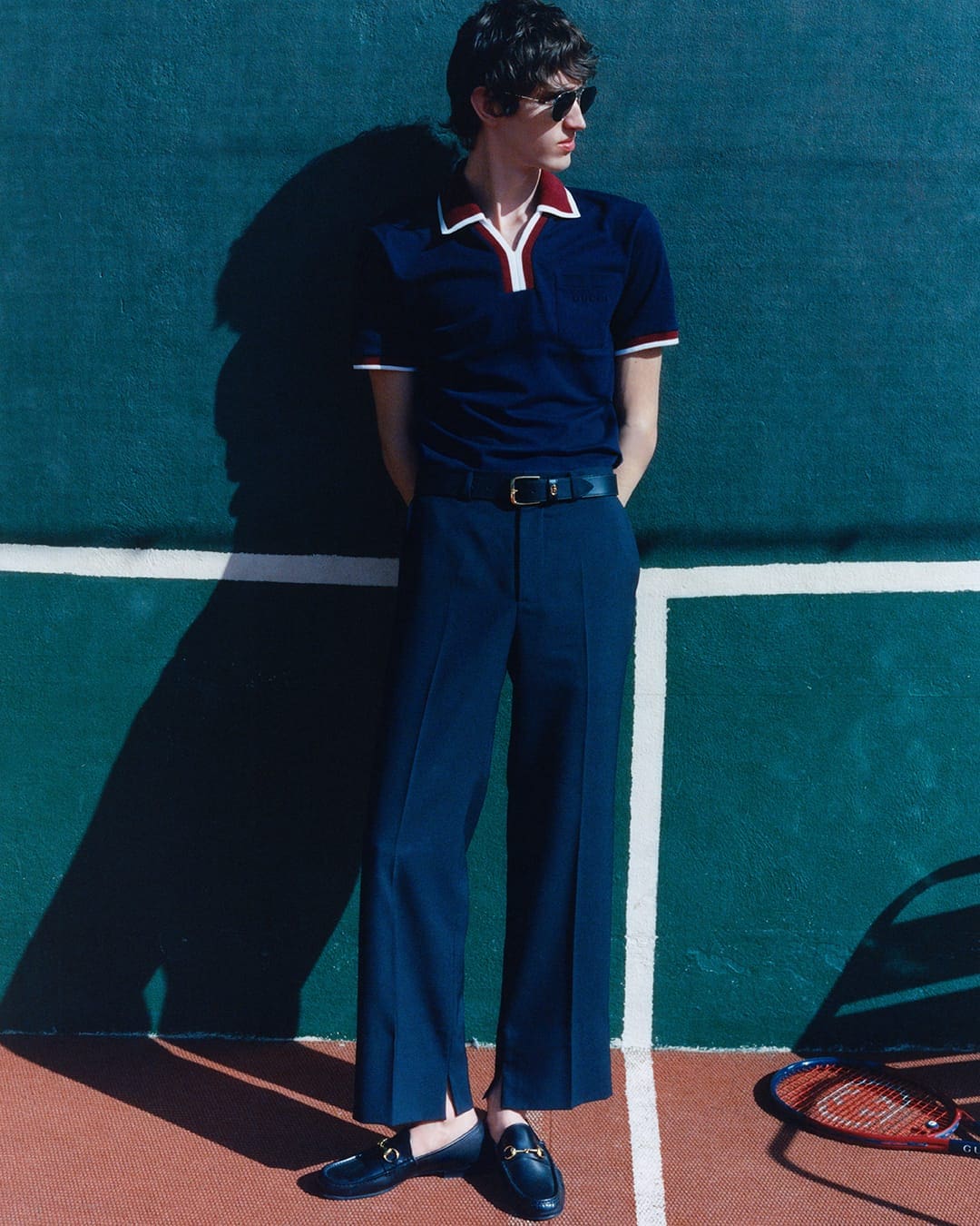 Man wearing a navy blue polo shirt and matching trousers standing against a green tennis court wall with white court lines.