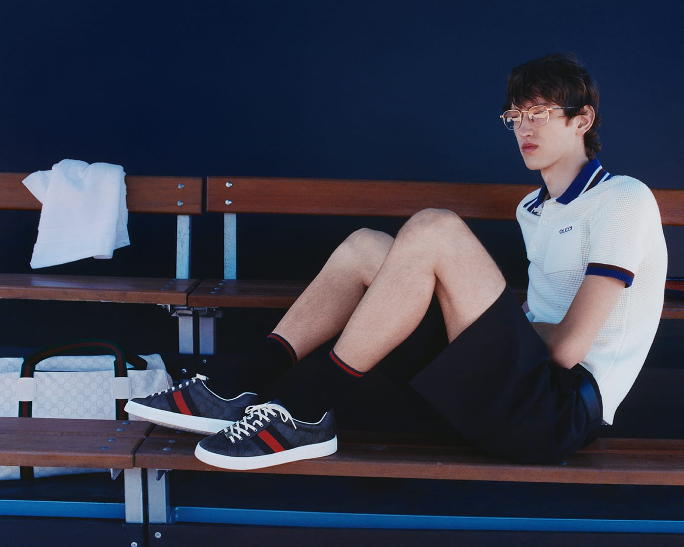 Young man wearing glasses, white polo shirt, and dark shorts seated on wooden stadium benches outdoors for Gucci ad campaign.