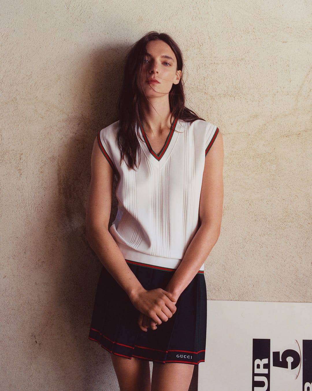 Model wearing a white V-neck vest and dark pleated skirt leaning against a textured wall for Gucci Tennis 2025 Ad Campaign.