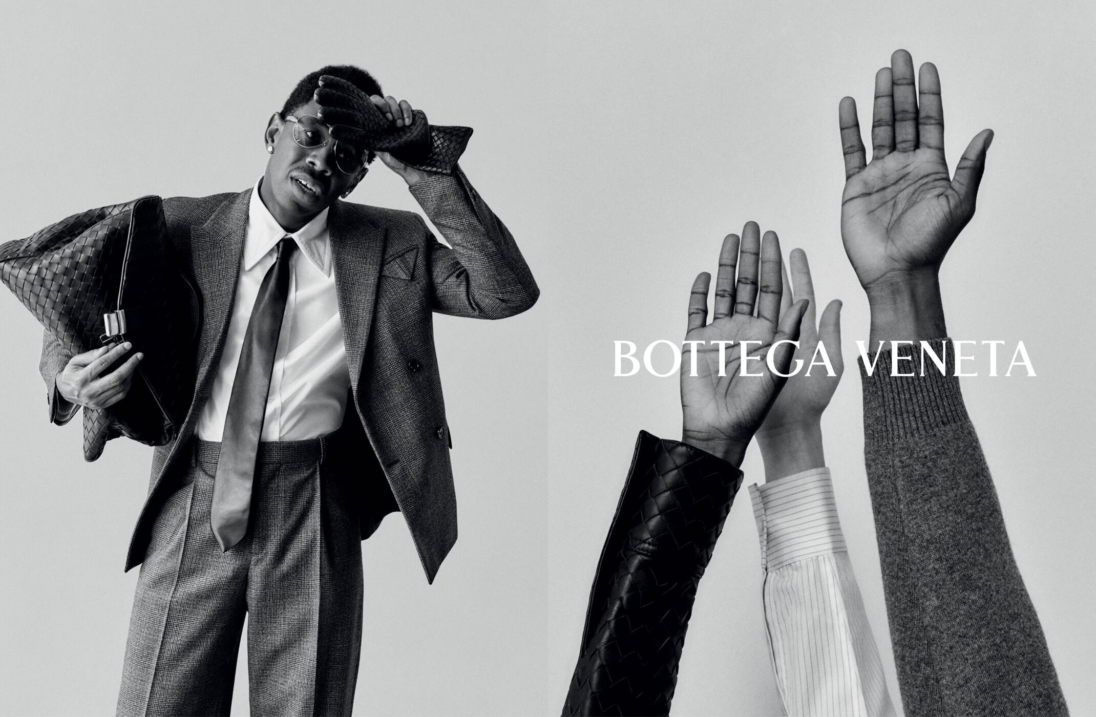 Black and white ad campaign image featuring a man in a grey suit holding a woven leather bag alongside raised hands.