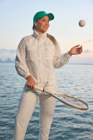 Donna Vekic wearing a white jacket and green cap holding a tennis racket near water for Mackage