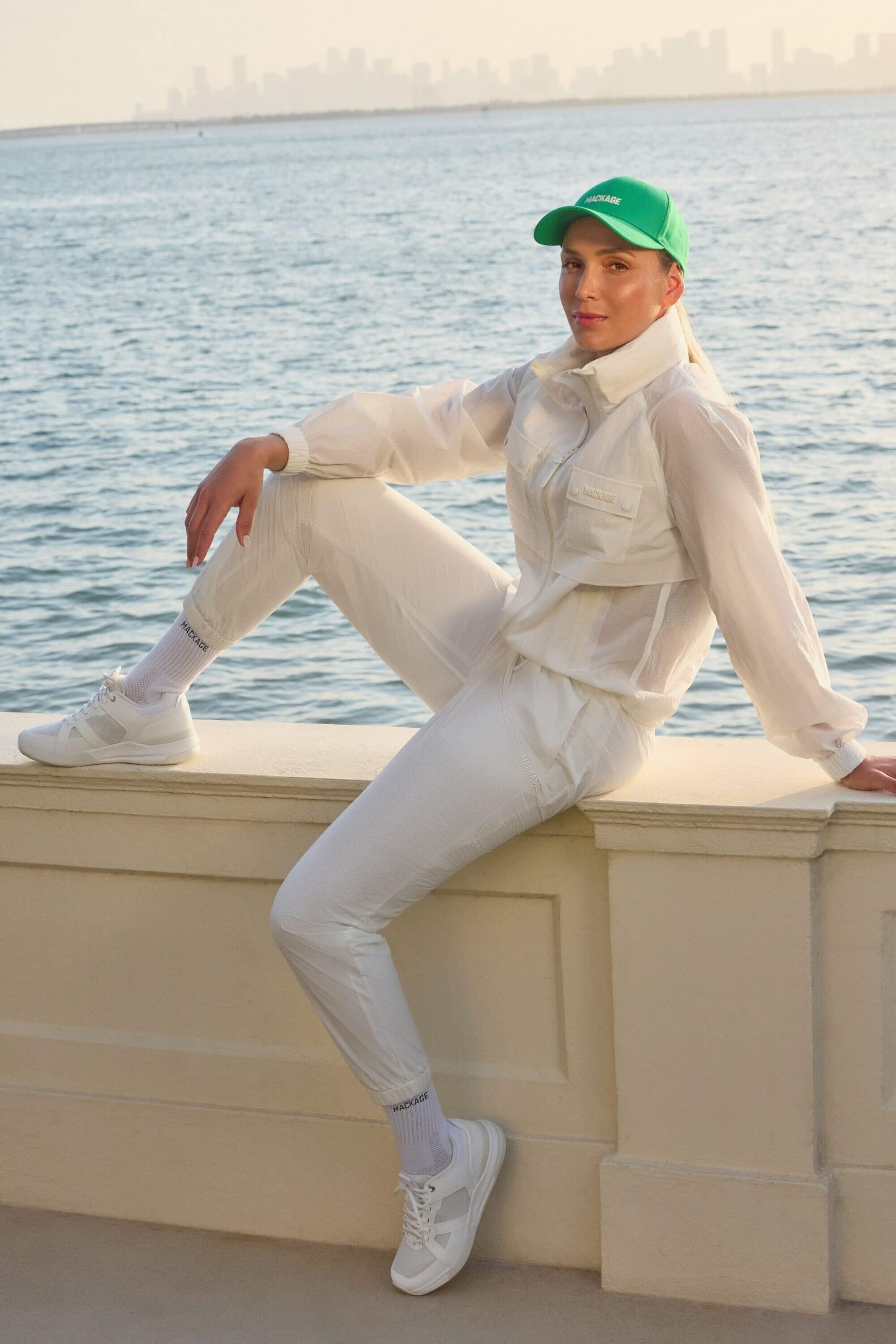 Model Donna Vekić wearing a green cap and white athletic outfit seated on a low wall against an ocean background.