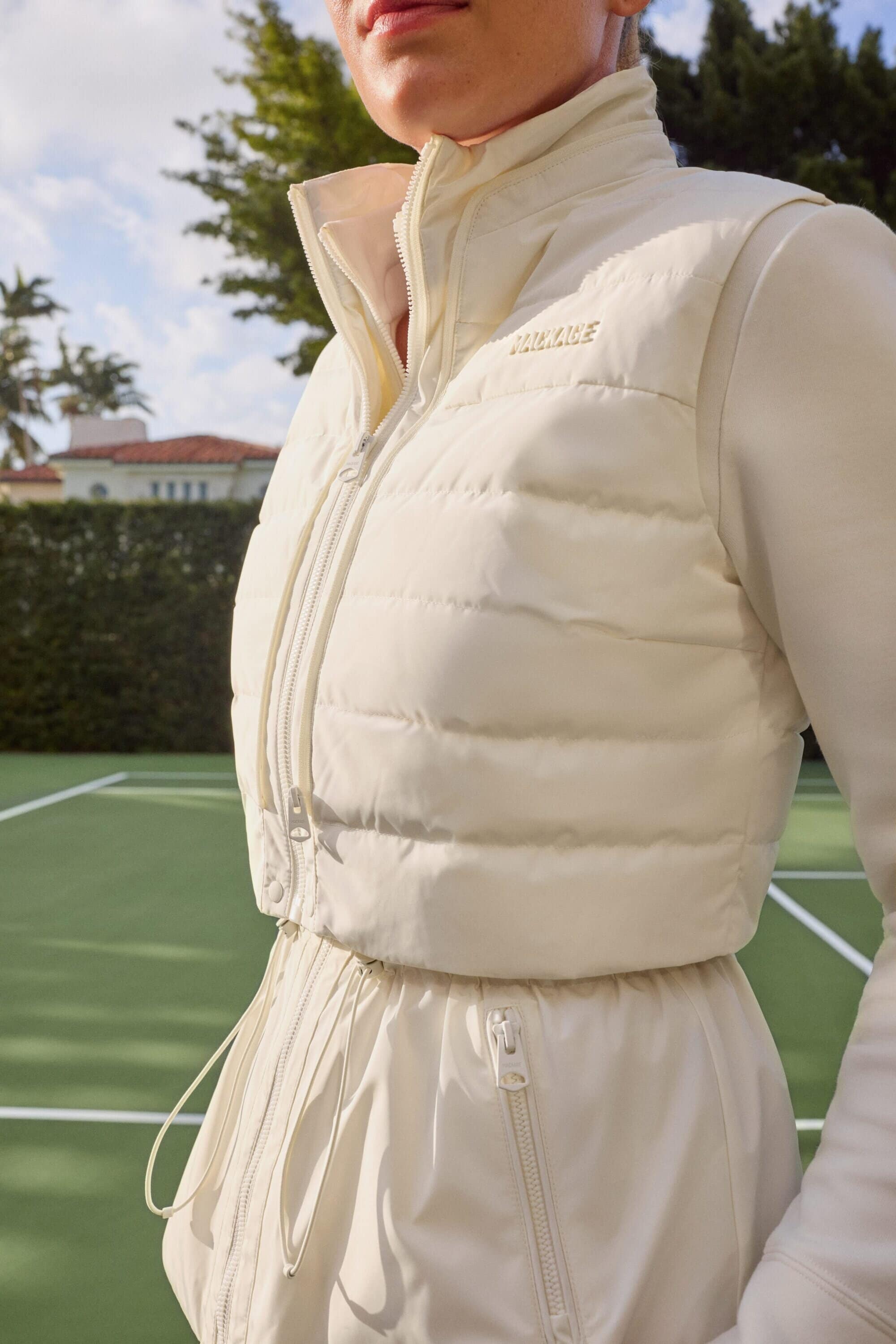 Model wearing a white padded vest and matching jacket standing on a tennis court outdoors under a clear sky.