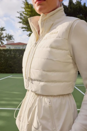 Model wearing a white padded vest and matching jacket standing on a tennis court outdoors under a clear sky.